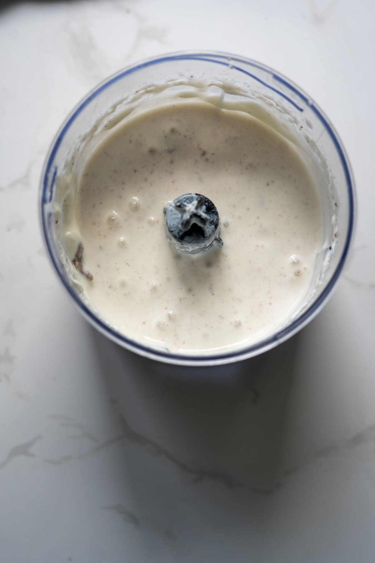 A top-down view of a blender jug containing a thick, creamy white mixture with small specks, set on a light-coloured surface.