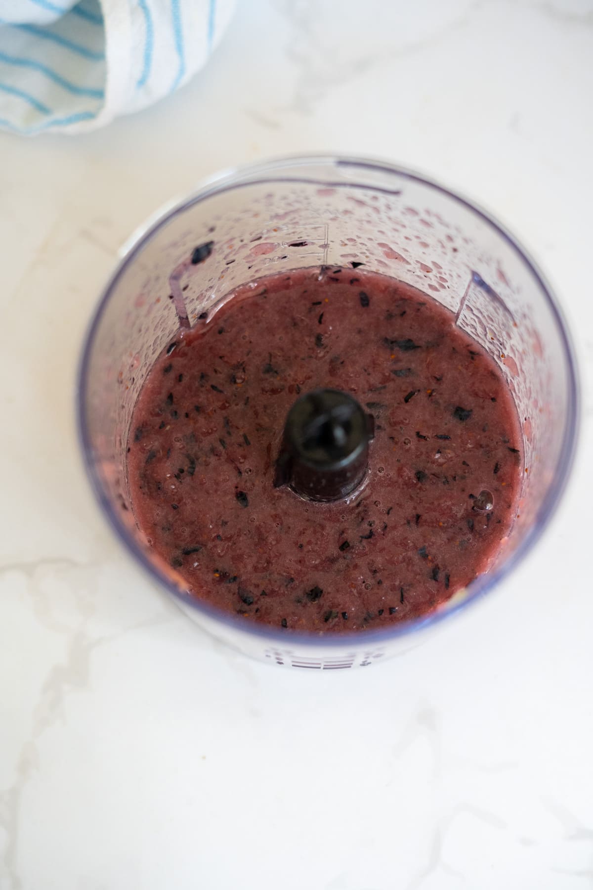 A blender cup containing a purple fruit smoothie mixture—reminiscent of the vibrant hues found in frozen cantaloupe blueberry dog treats—sits on a white worktop, with a striped tea towel in the background.