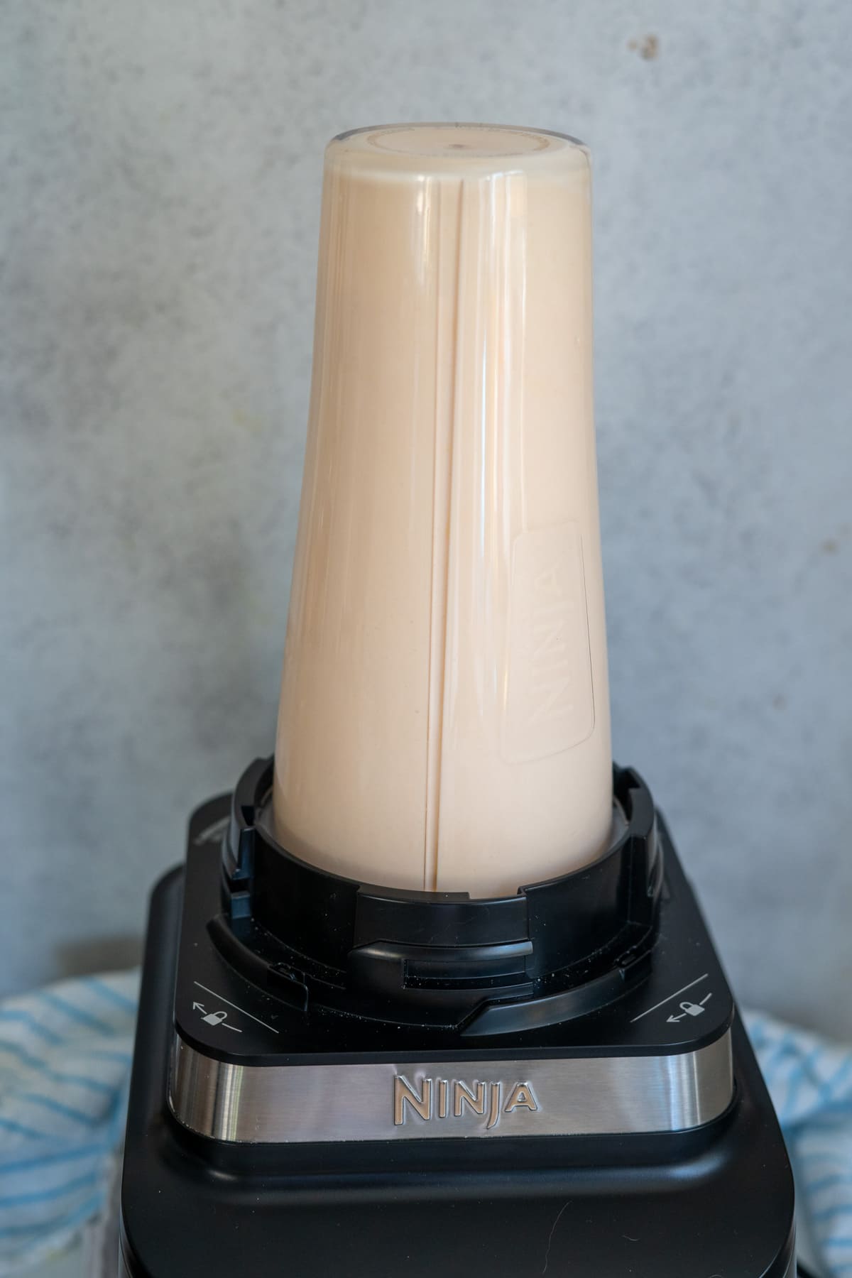 A Ninja blender with a cup attached upside down, filled with a light beige smoothie, sits on a worktop against a grey background.
