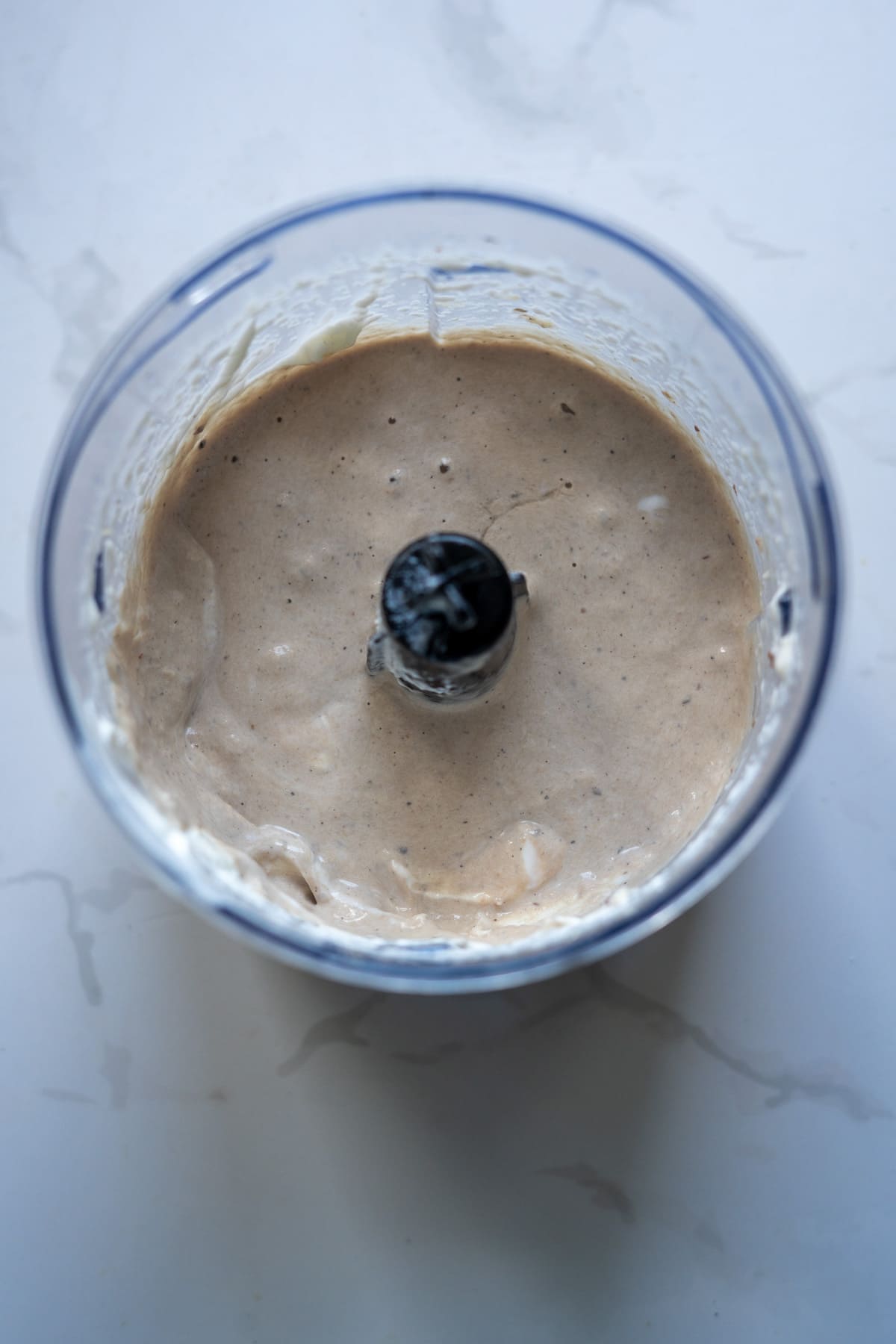 A food processor bowl containing a thick, creamy beige sardine ice cream mixture viewed from above on a light surface.