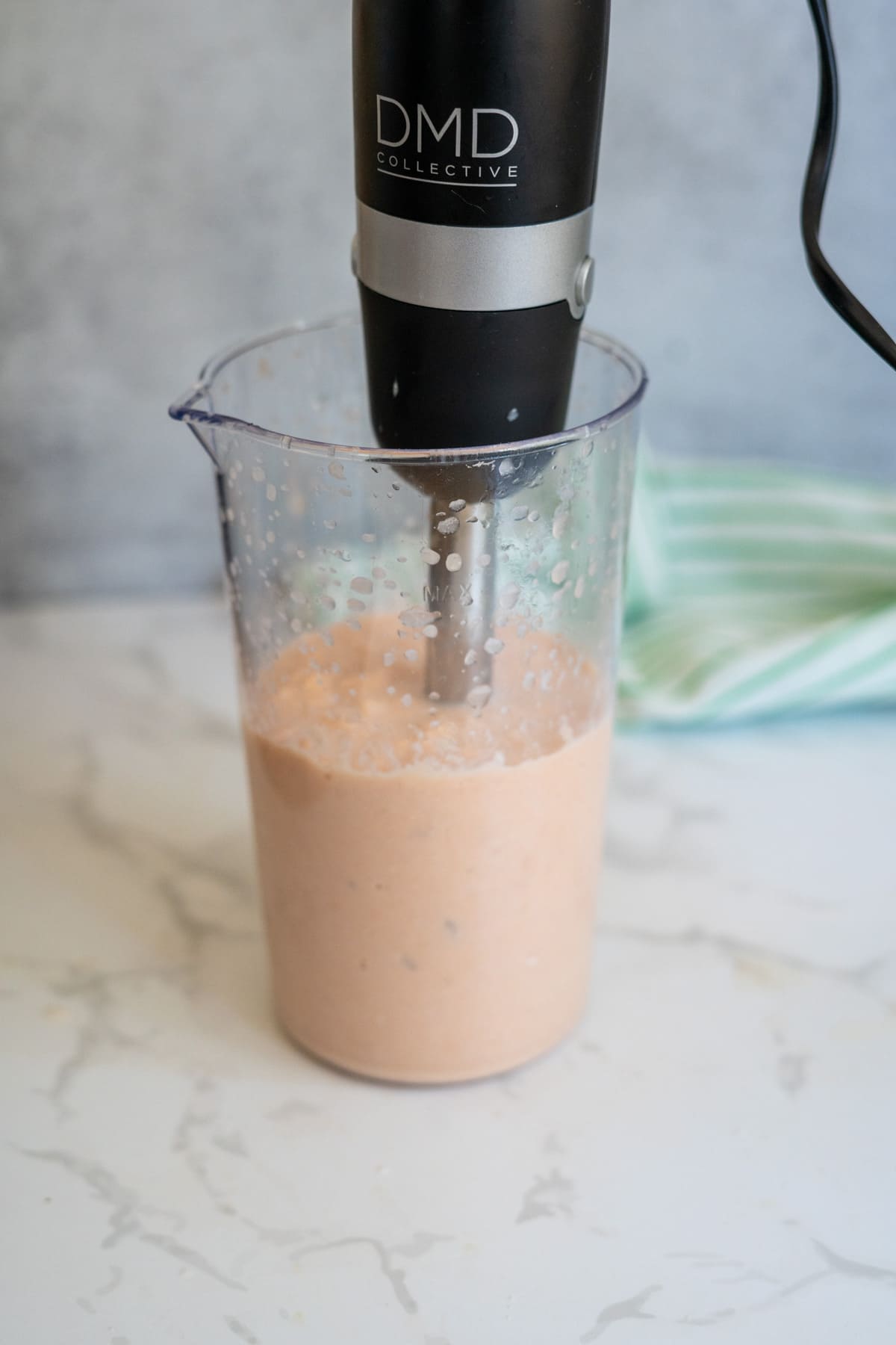 An immersion blender is blending a light pink mixture, perfect for making salmon bone broth gummies, in a clear plastic container on a white worktop.
