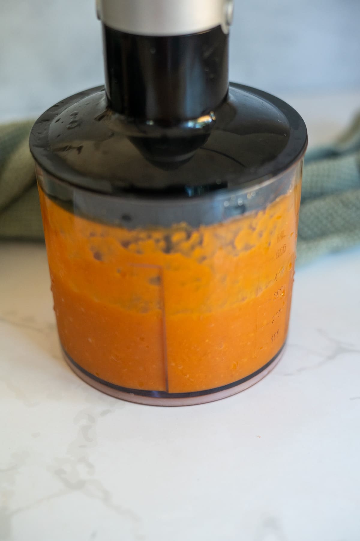 Clear container with orange sauce being blended, placed on a white worktop, with a green kitchen towel in the background—perfect for making Cantaloupe Pumpkin Frozen Dog Treats.