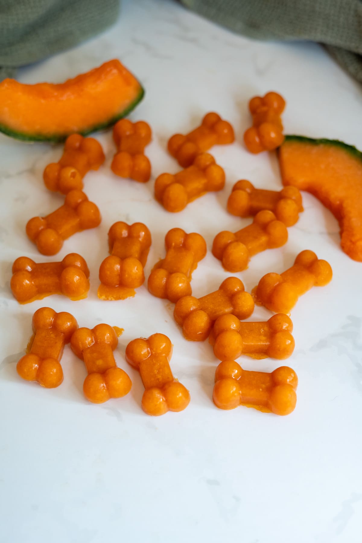 Small, orange, bone-shaped Cantaloupe Pumpkin Frozen Dog Treats are arranged on a white surface next to sliced cantaloupe.