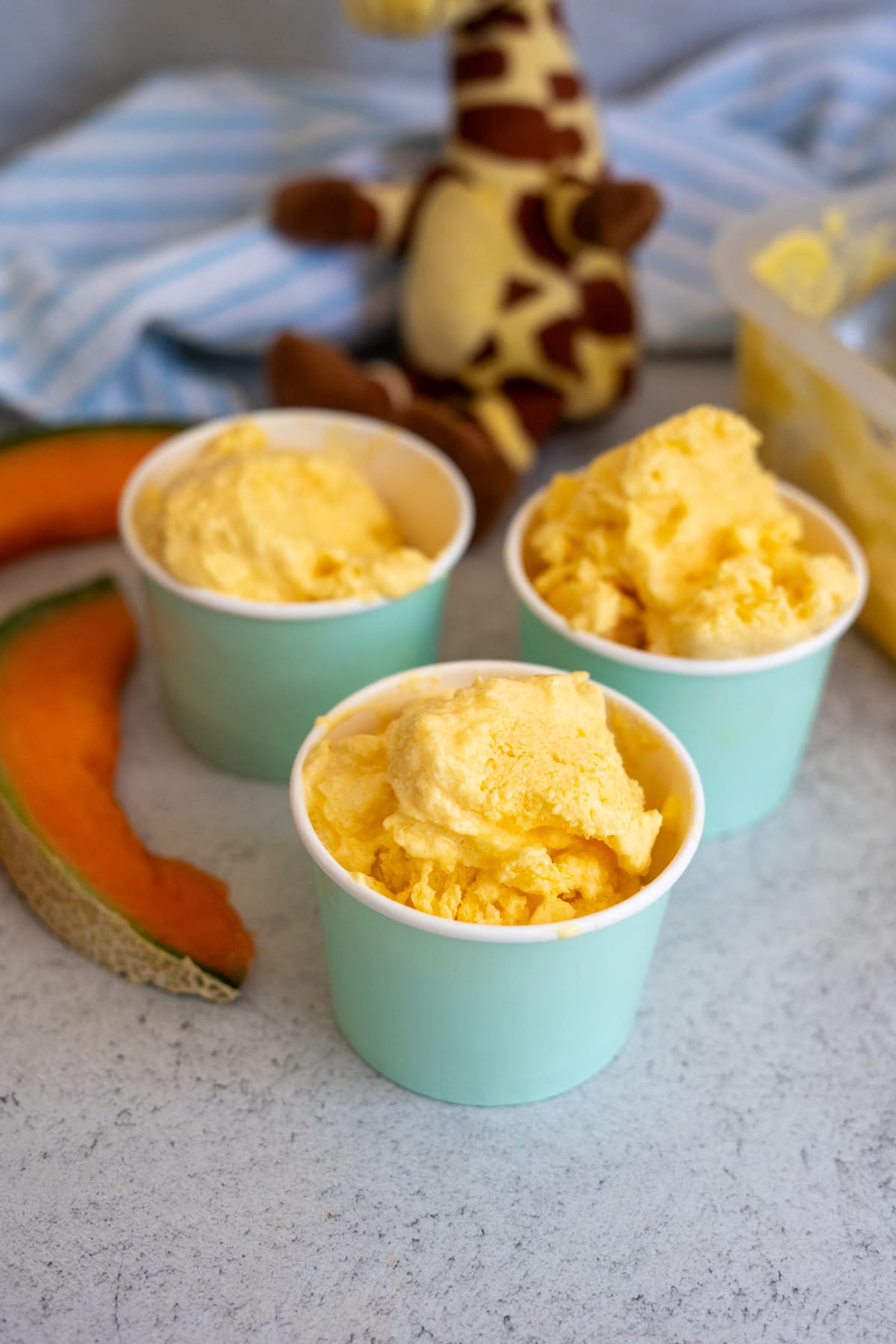 Three cups of orange sorbet are placed on a worktop with cantaloupe slices nearby. A plush giraffe toy and a striped cloth are in the background.