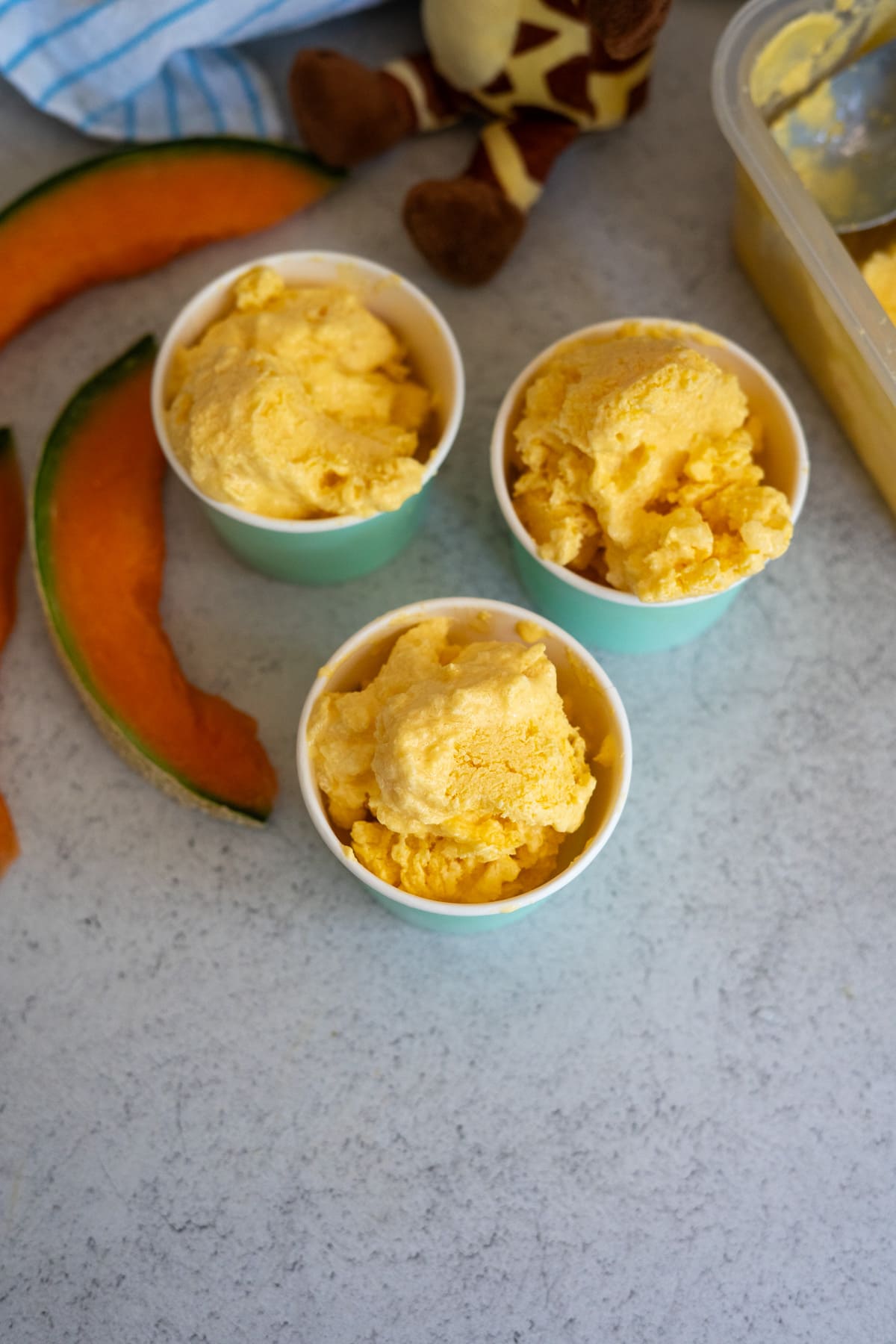 Three cups of orange melon ice cream sit on a worktop next to cantaloupe slices, a stuffed giraffe toy, and a tub of extra ice cream.