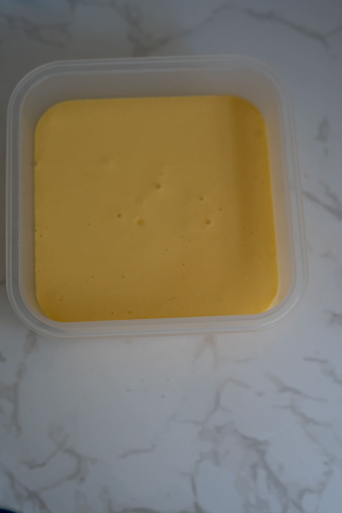 A square plastic container filled with smooth, yellow liquid or batter sits on a white marbled surface.