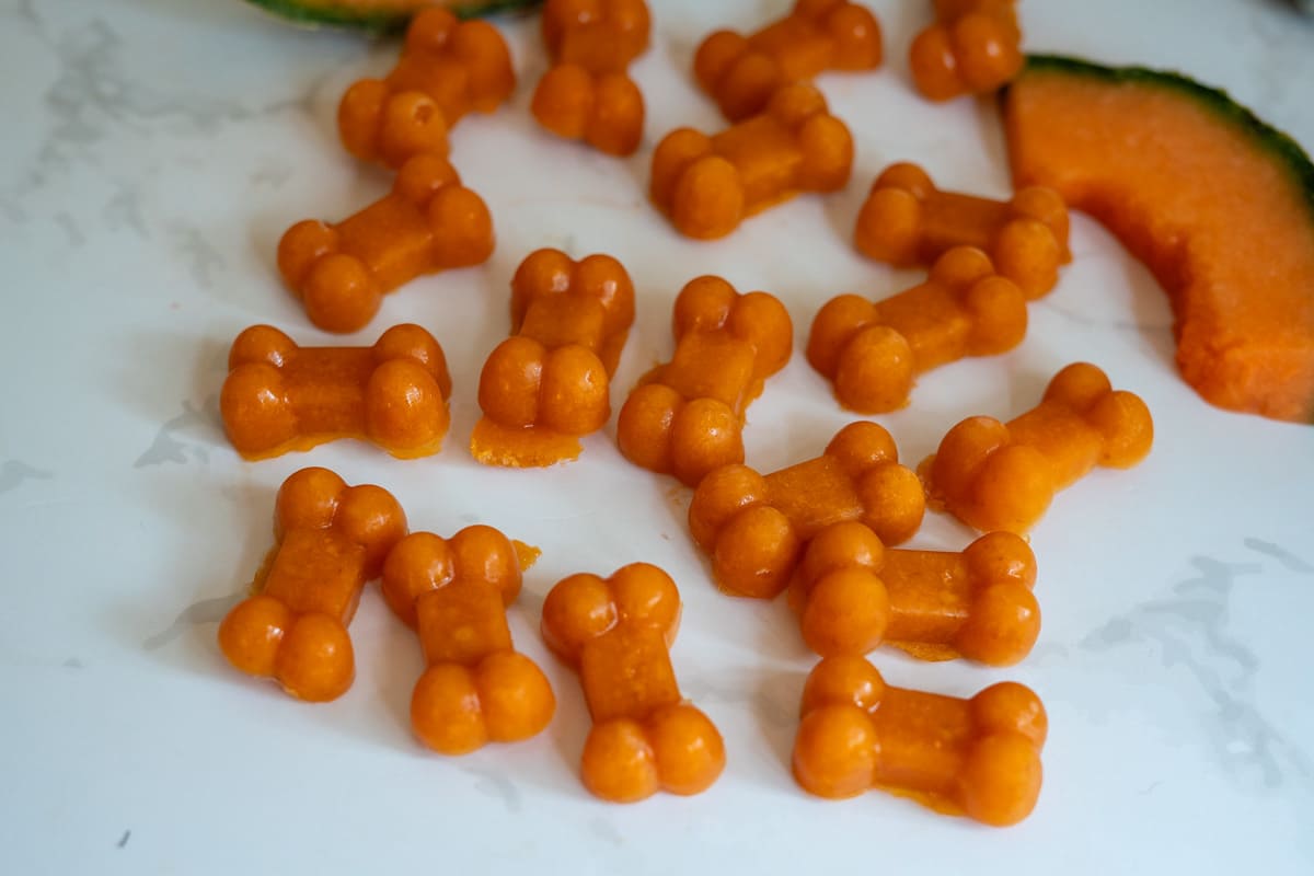 Small, orange, bone-shaped Cantaloupe Pumpkin Frozen Dog Treats are scattered on a white surface with slices of cantaloupe melon in the background.