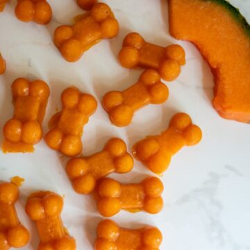 Small, orange, bone-shaped treats are arranged on a white surface next to a slice of melon, perfectly capturing the refreshing appeal of Cantaloupe Pumpkin Frozen Dog Treats.
