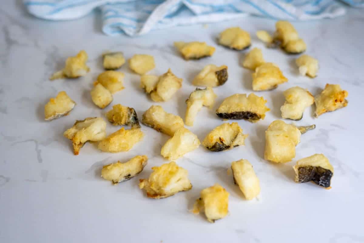 Small pieces of fried battered cod bites for dogs are spread out on a white surface with a blue and white cloth in the background.