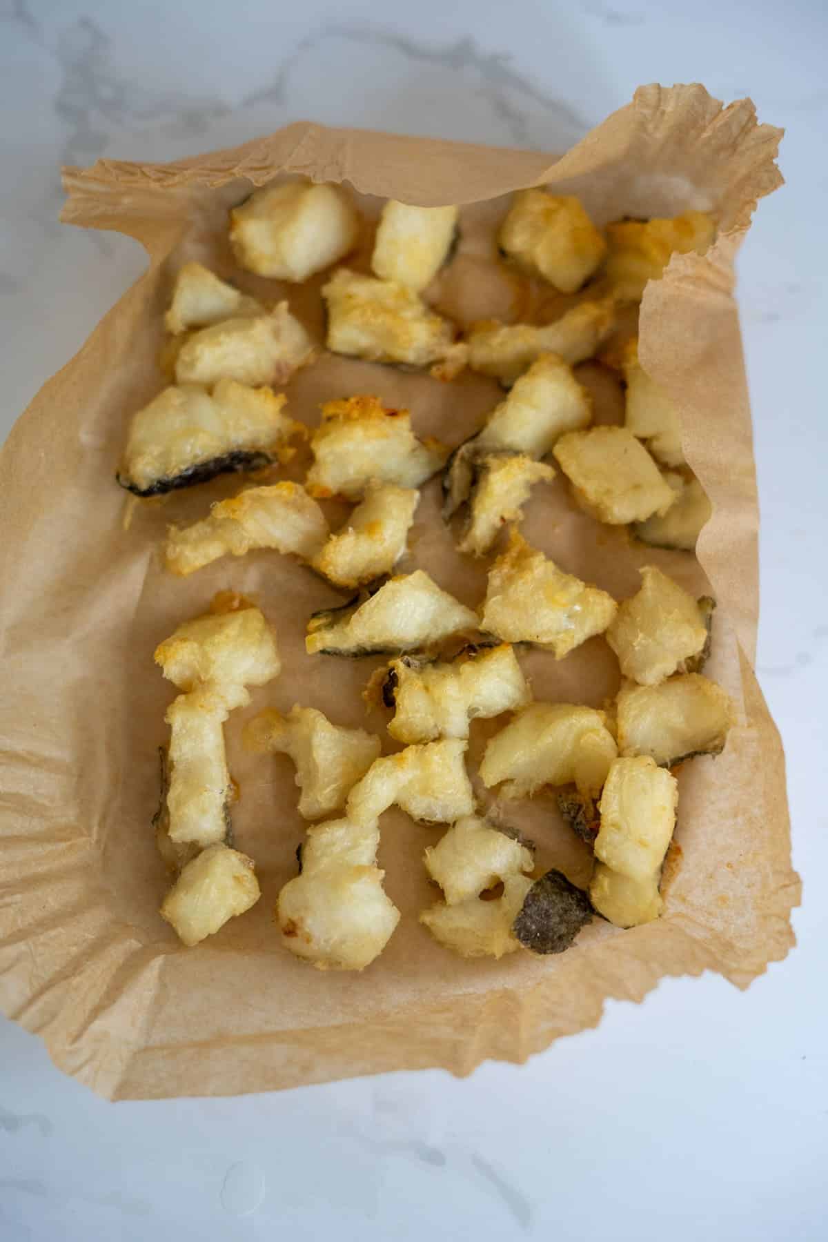 Battered and fried fish pieces, resembling savoury cod bites for dogs, are arranged on a sheet of brown baking parchment atop a white surface.