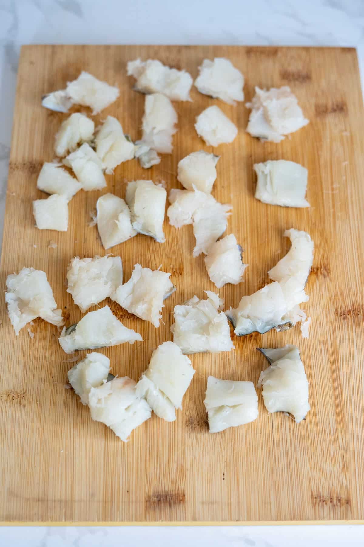 Pieces of raw white fish, like cod bites for dogs, are cut into chunks and spread out on a wooden chopping board.