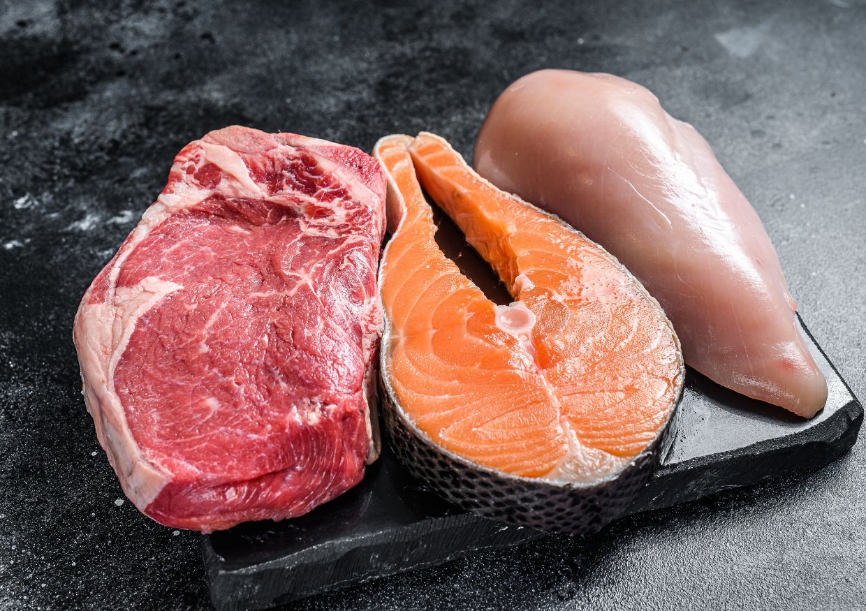 Raw beef steak, salmon fillet, and chicken breast are arranged side by side on a black slate surface.