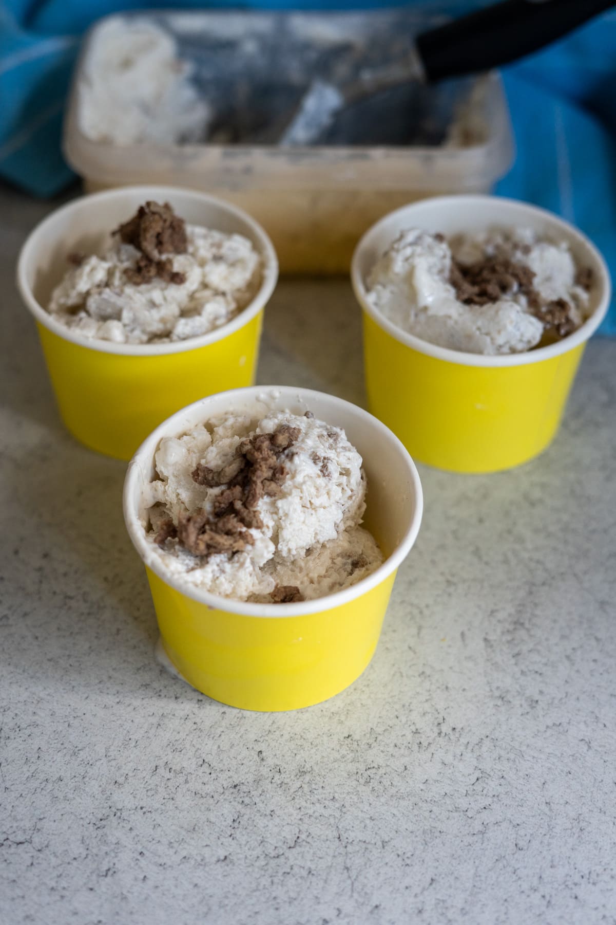 Three yellow cups filled with scoops of cookies and cream ice cream are placed on a worktop, alongside a tub of beef dog ice cream and an ice cream scoop in the background.