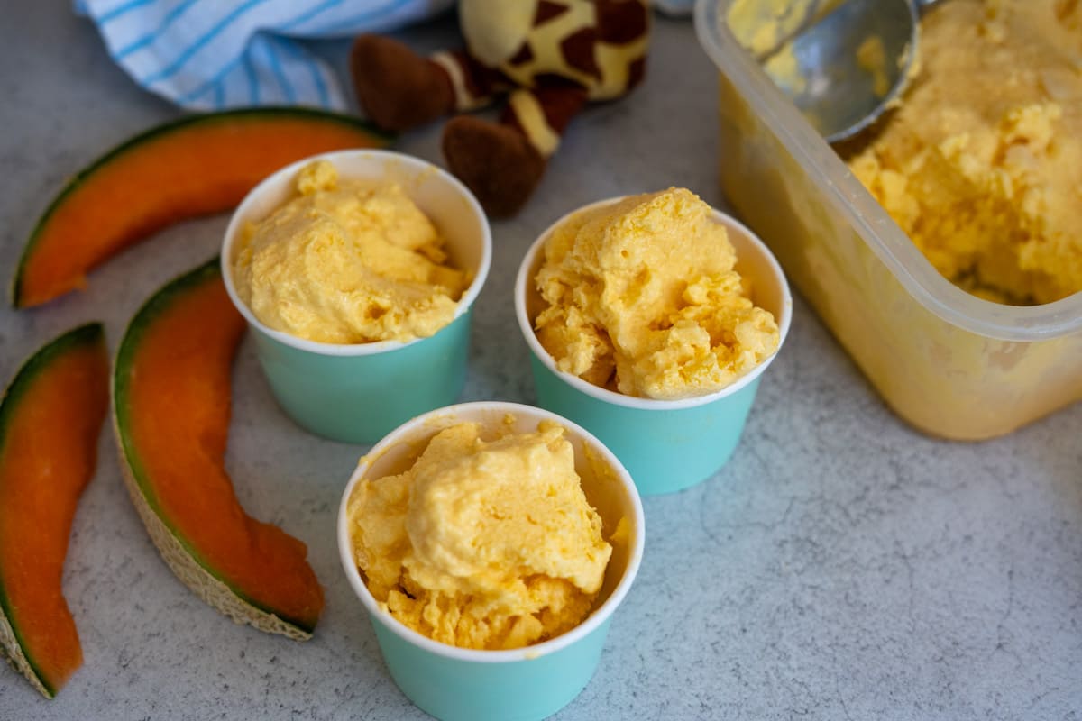 Three blue cups filled with orange ice cream sit next to a tub of ice cream and three cantaloupe slices on a worktop.