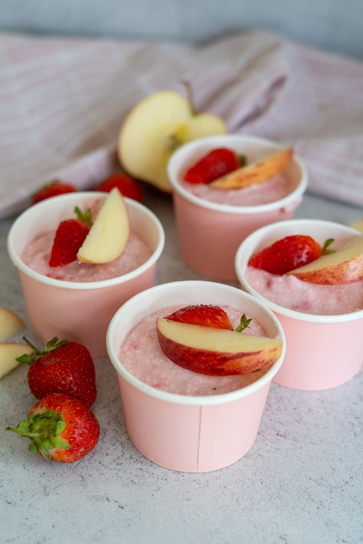 Four pink cups filled with a pink dessert, each topped with a strawberry and an apple slice, resemble the perfect treat for a summer day—much like apple strawberry ice cream—whilst fresh fruit and a striped cloth set a cheerful background.