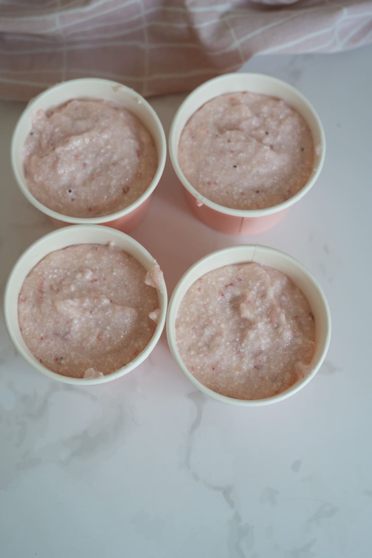 Four cups filled with a pink, creamy apple and strawberry mixture are arranged on a white surface with a pink cloth in the background.