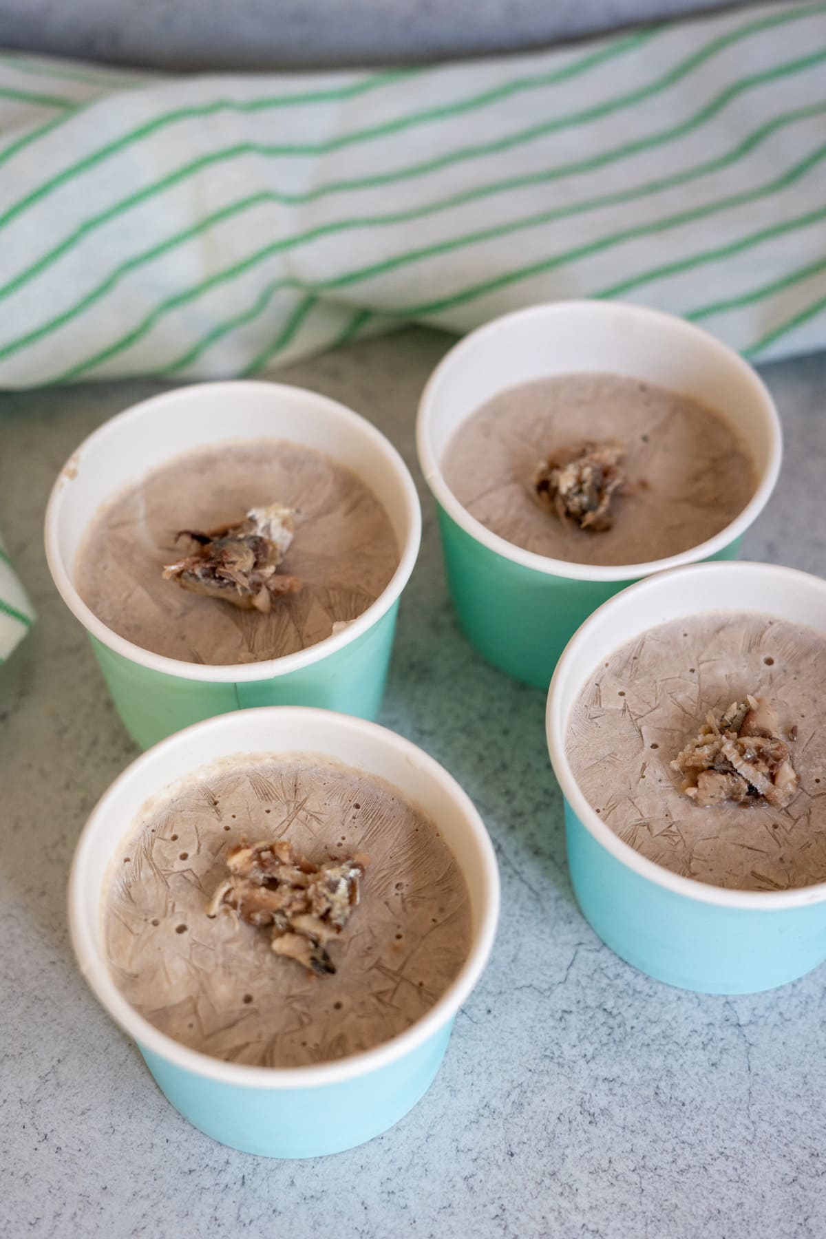 Four small blue paper cups filled with brown sardine ice cream, each topped with a small cluster of nuts or granola, placed on a light worktop with a striped cloth in the background.