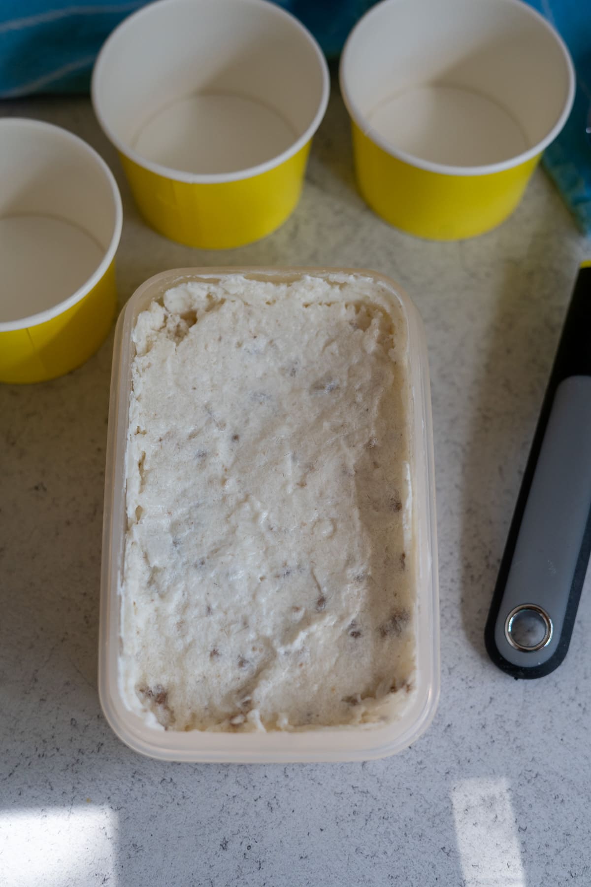 A rectangular container filled with a creamy, textured mixture sits on a worktop next to three empty yellow paper cups and an ice cream scoop.