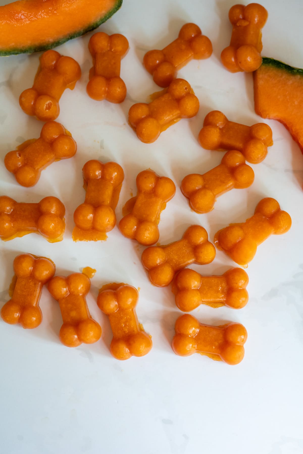 Small, orange, bone-shaped Cantaloupe Pumpkin Frozen Dog Treats are arranged on a white surface with slices of cantaloupe visible in the background.