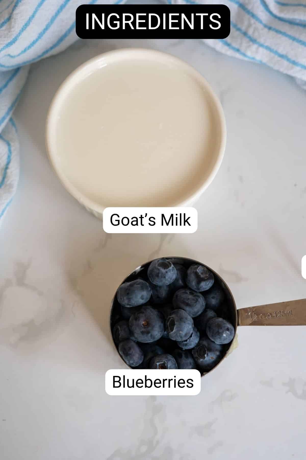 A measuring jug of blueberries and a small bowl of goat’s milk sit on a white surface with a striped tea towel in the background, ready to be combined into delicious Frozen Goat Milk Dog Treats.