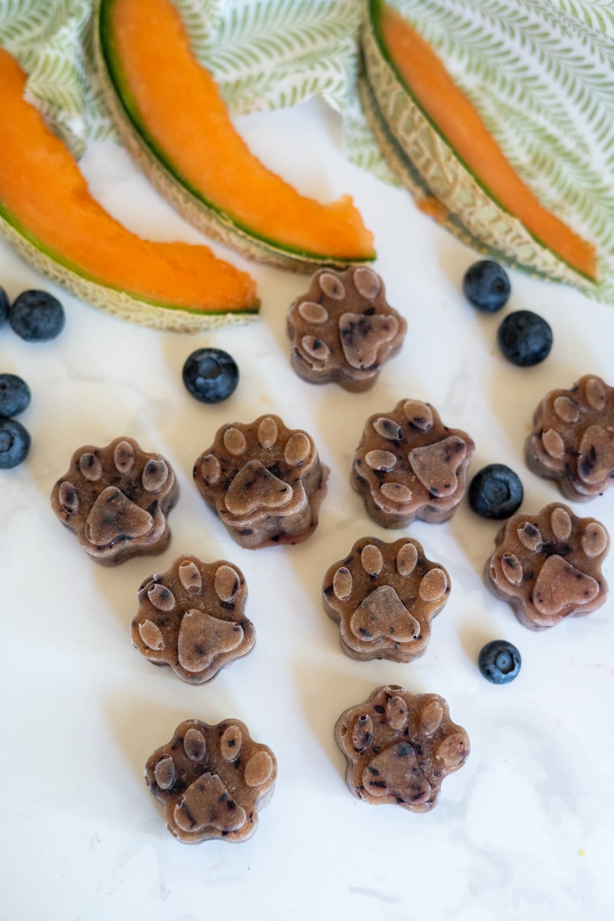 Frozen cantaloupe blueberry dog treats shaped like paw prints are arranged on a white surface with fresh blueberries and cantaloupe slices, near a green and white patterned cloth.