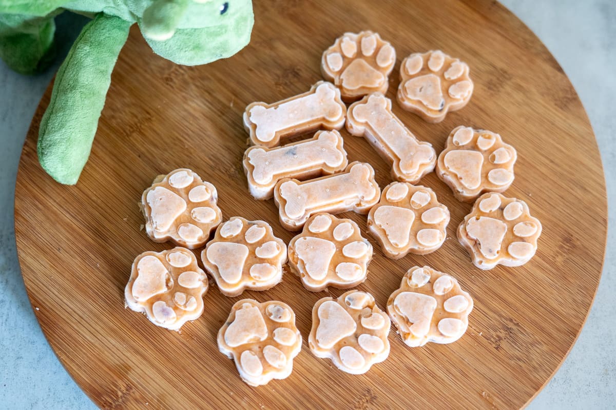 Savoury frozen dog treats shaped like bones and paw prints are arranged on a round wooden board, with part of a green plush toy visible in the corner.