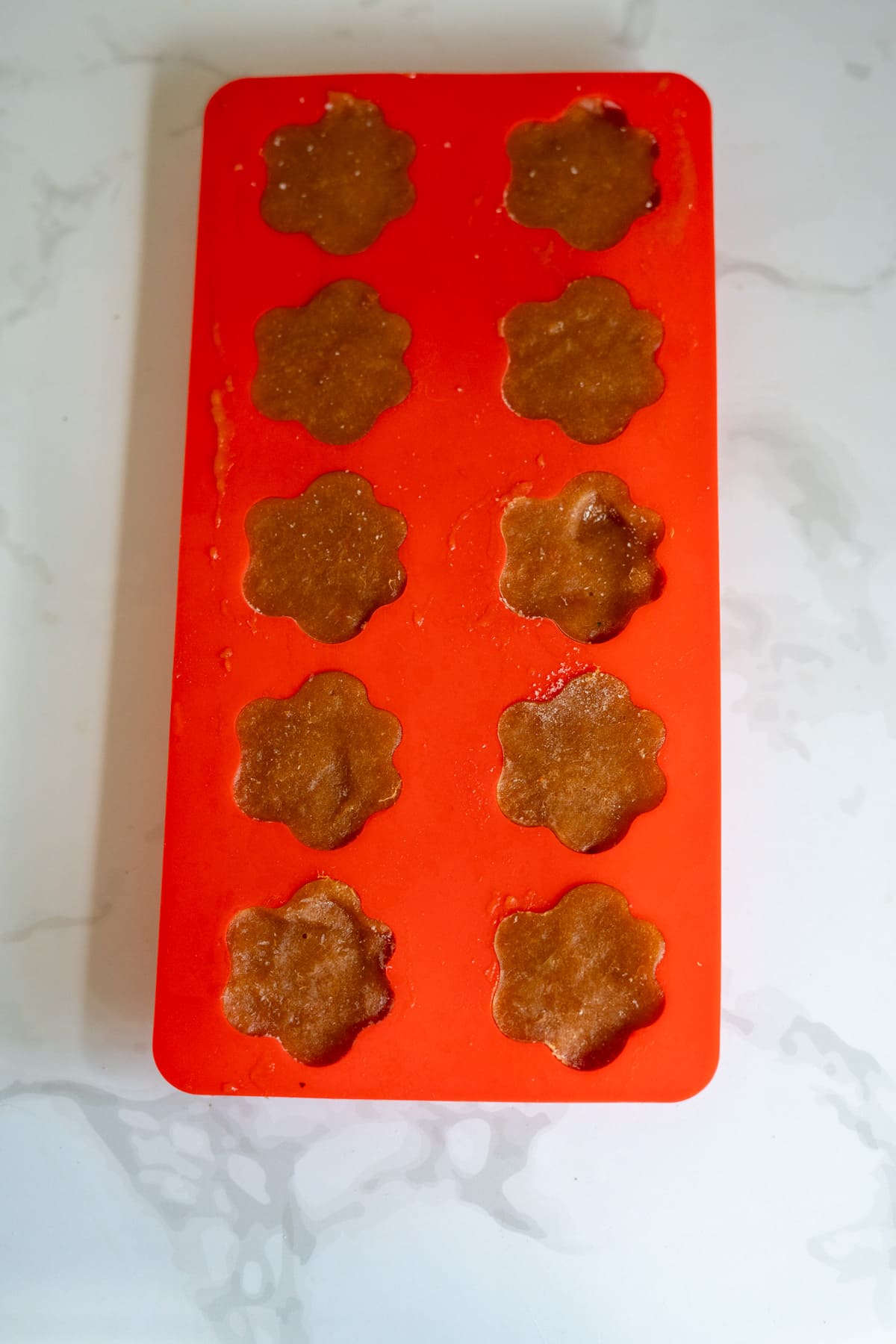 A red silicone mould with 12 flower-shaped cavities is filled with brown batter, placed on a white marble surface.