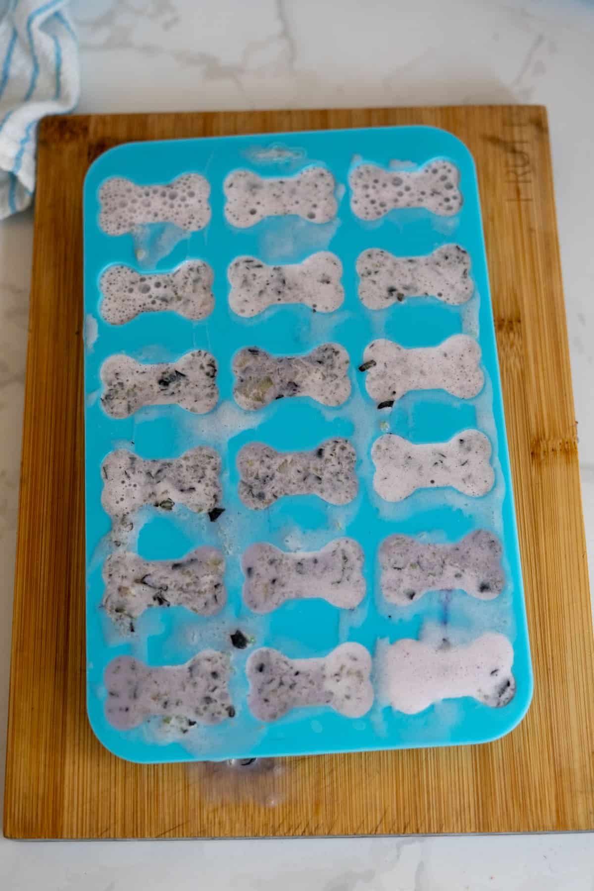 A blue silicone mould shaped like dog bones is filled with a speckled mixture of Frozen Goat Milk Dog Treats and placed on a wooden chopping board.