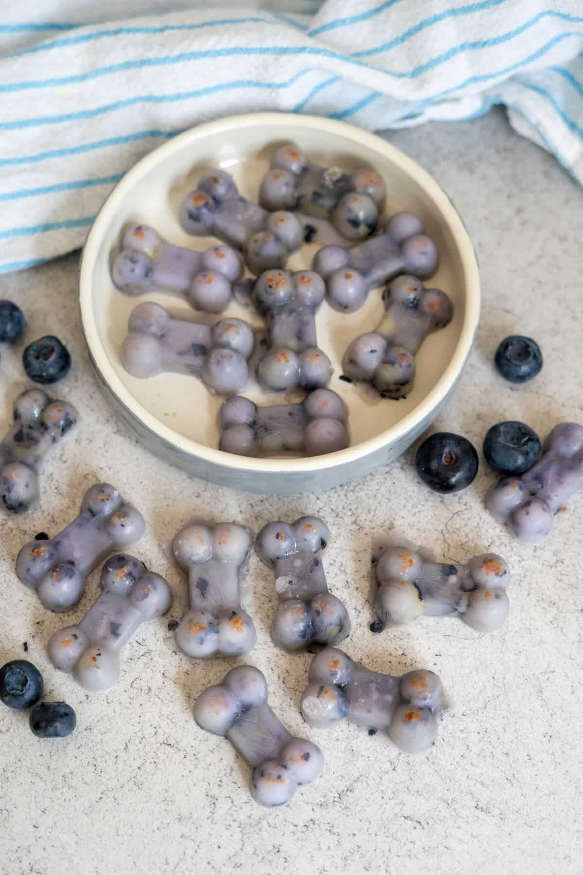 A bowl and surface with bone-shaped Frozen Goat's Milk Dog Treats containing blueberries, next to a few loose blueberries and a blue-striped cloth.
