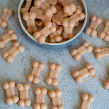 A bowl filled with bone-shaped treats is surrounded by more of the same treats scattered on a light grey surface.