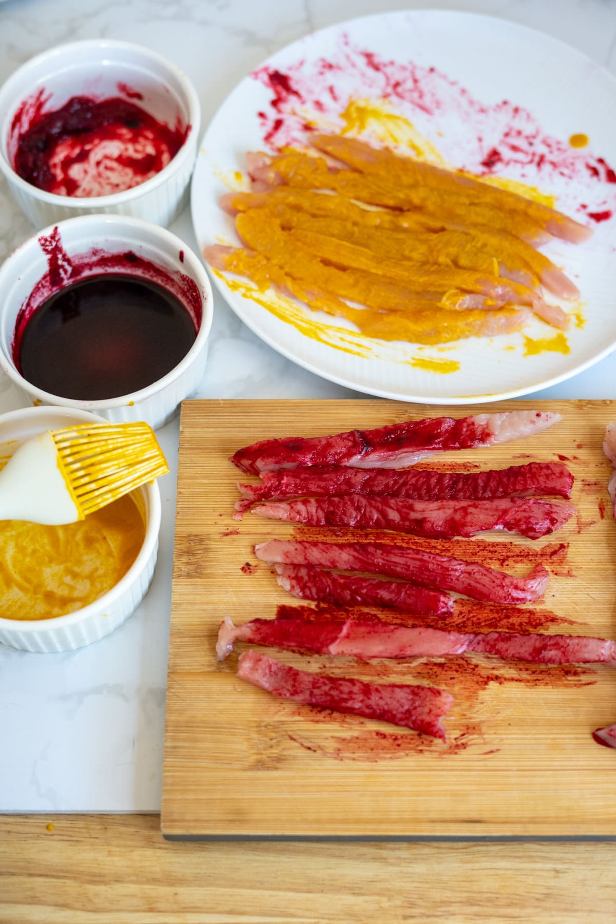 Slices of fish being marinated with red and yellow seasonings on a chopping board and plate, with small bowls of sauces, a brush nearby, and turkey jerky for dogs placed alongside, ready for a flavourful preparation.
