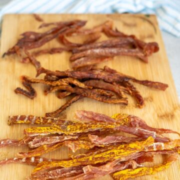 Three piles of homemade beef jerky strips, each seasoned differently, arranged on a wooden chopping board with a blue and white cloth in the background.