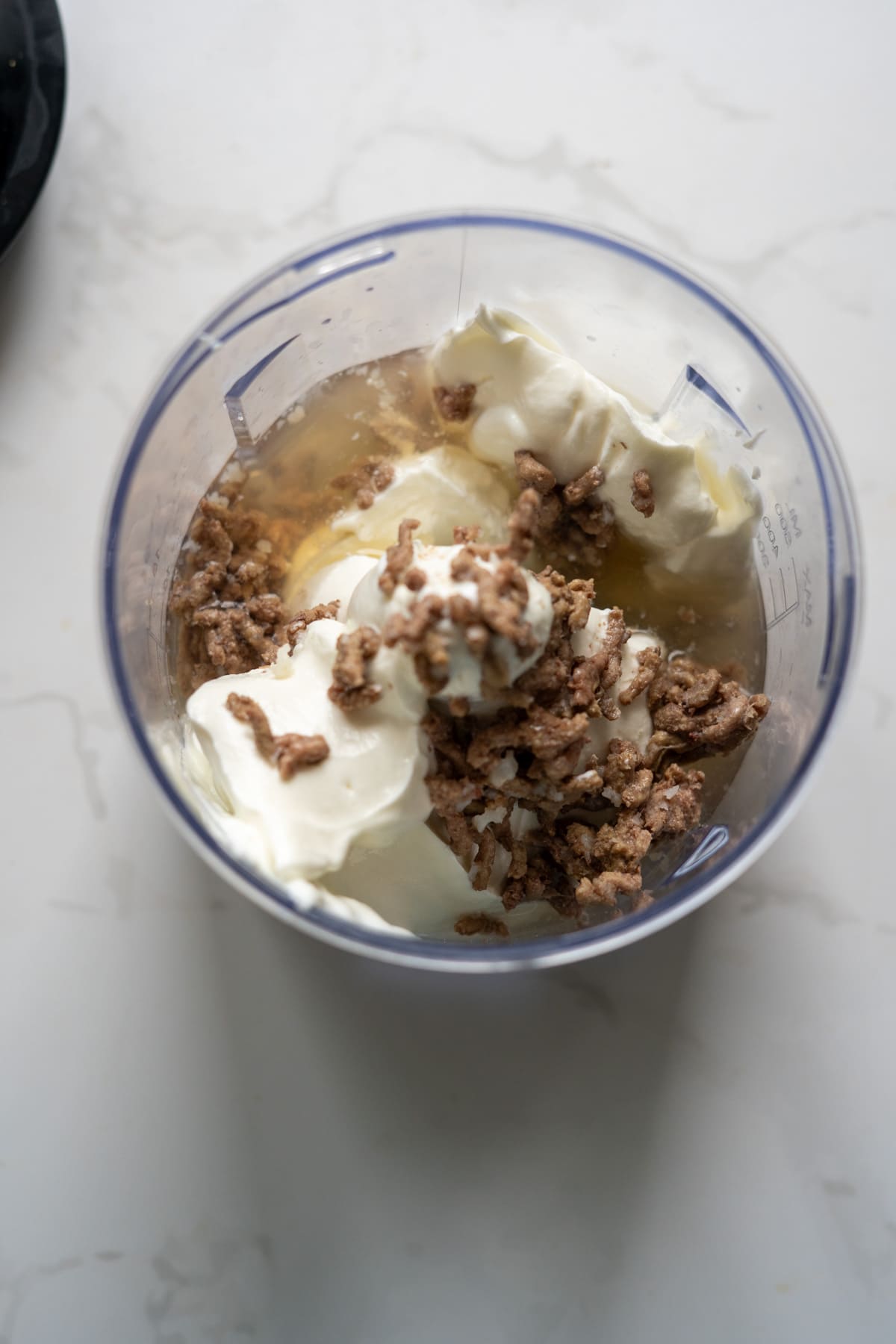 A blender jug containing minced meat, soured cream, and a light-coloured liquid on a white worktop.