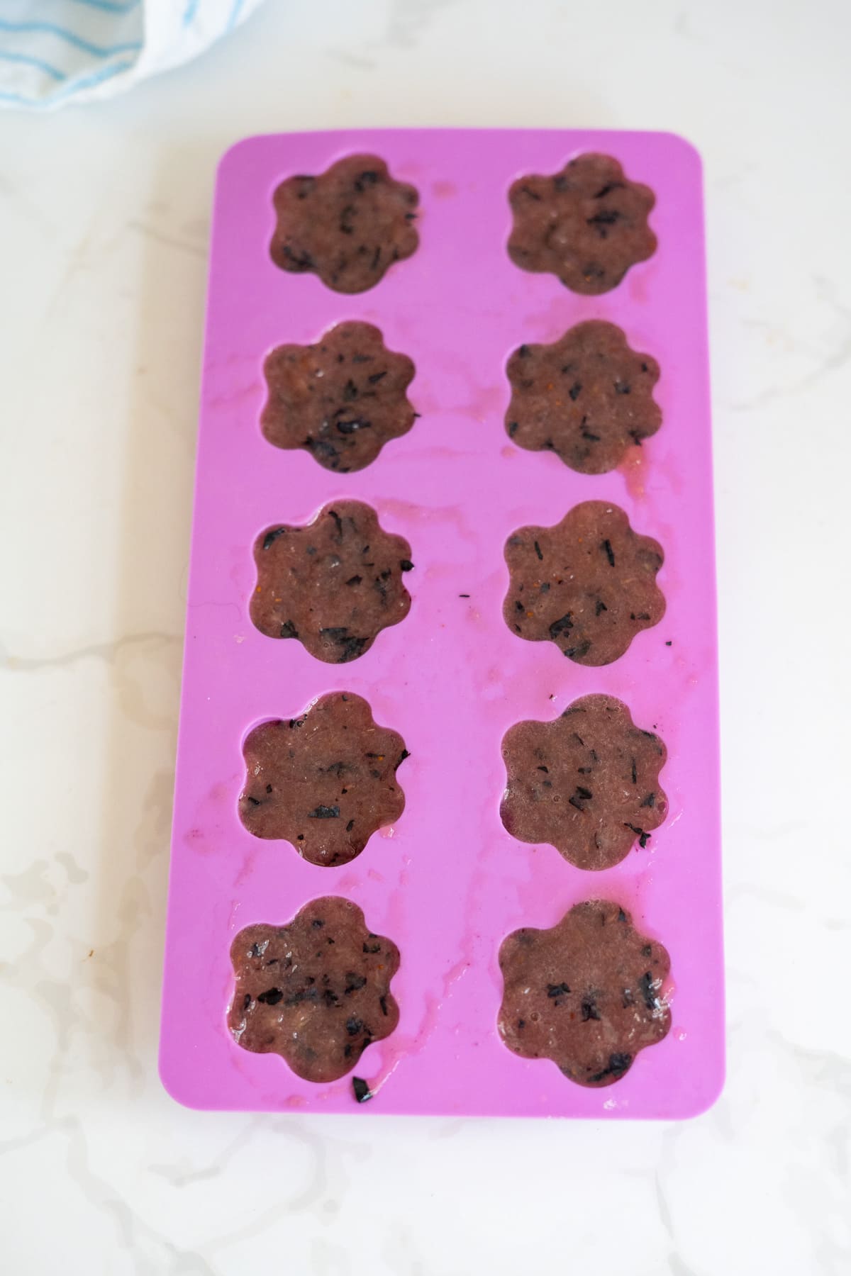 A pink silicone mould tray holds twelve flower-shaped portions of berry-filled batter, perfect for making frozen cantaloupe blueberry dog treats, resting on a white countertop.