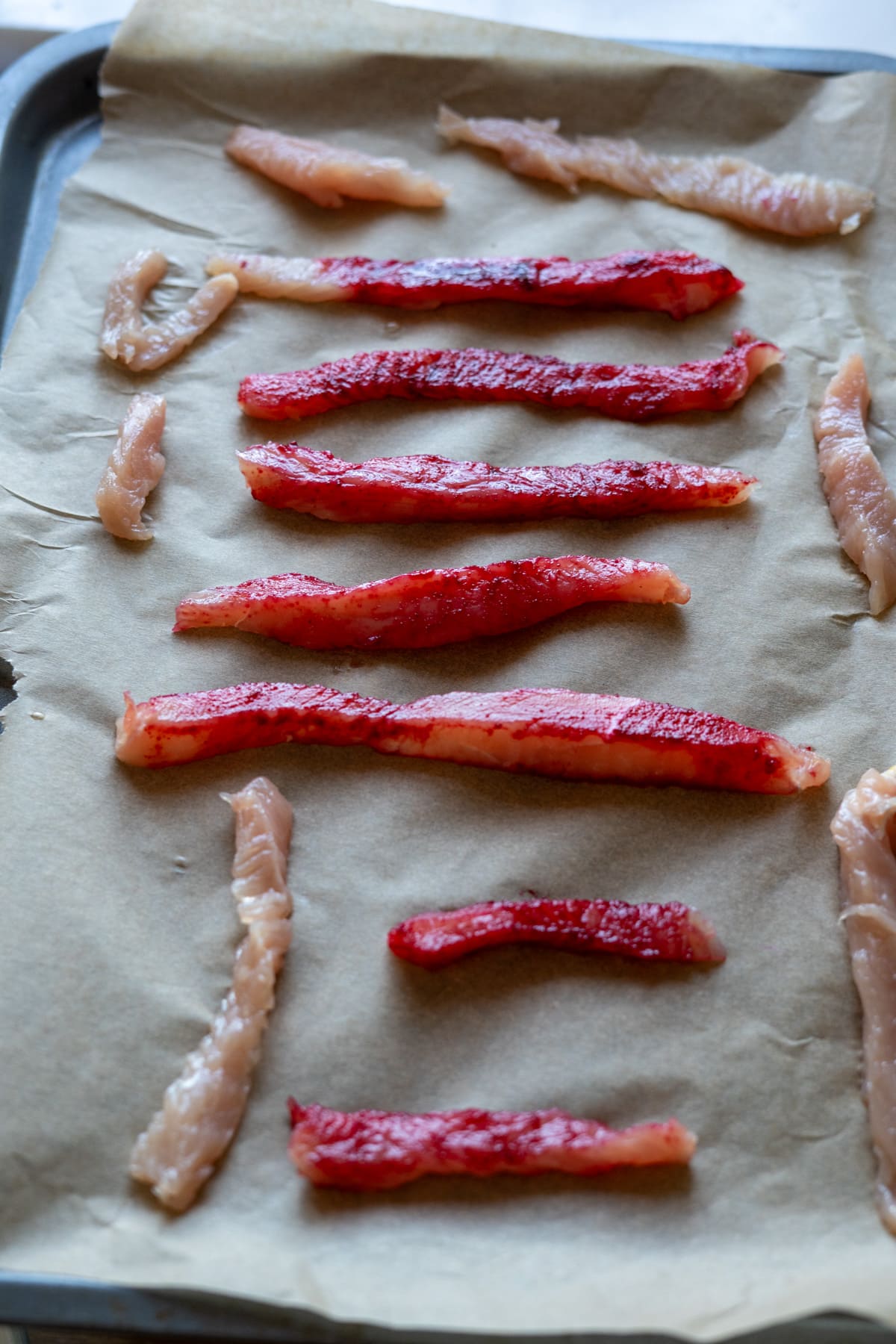 Raw strips of light and red fish fillets, reminiscent of turkey jerky for dogs, arranged on a baking tray lined with baking parchment.