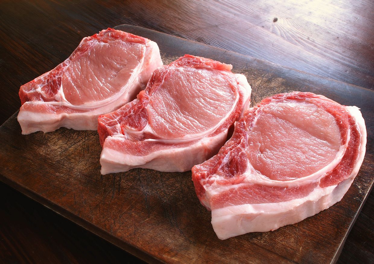 Three raw bone-in pork chops, considered by many as the best protein for dogs, are arranged side by side on a dark wooden chopping board.
