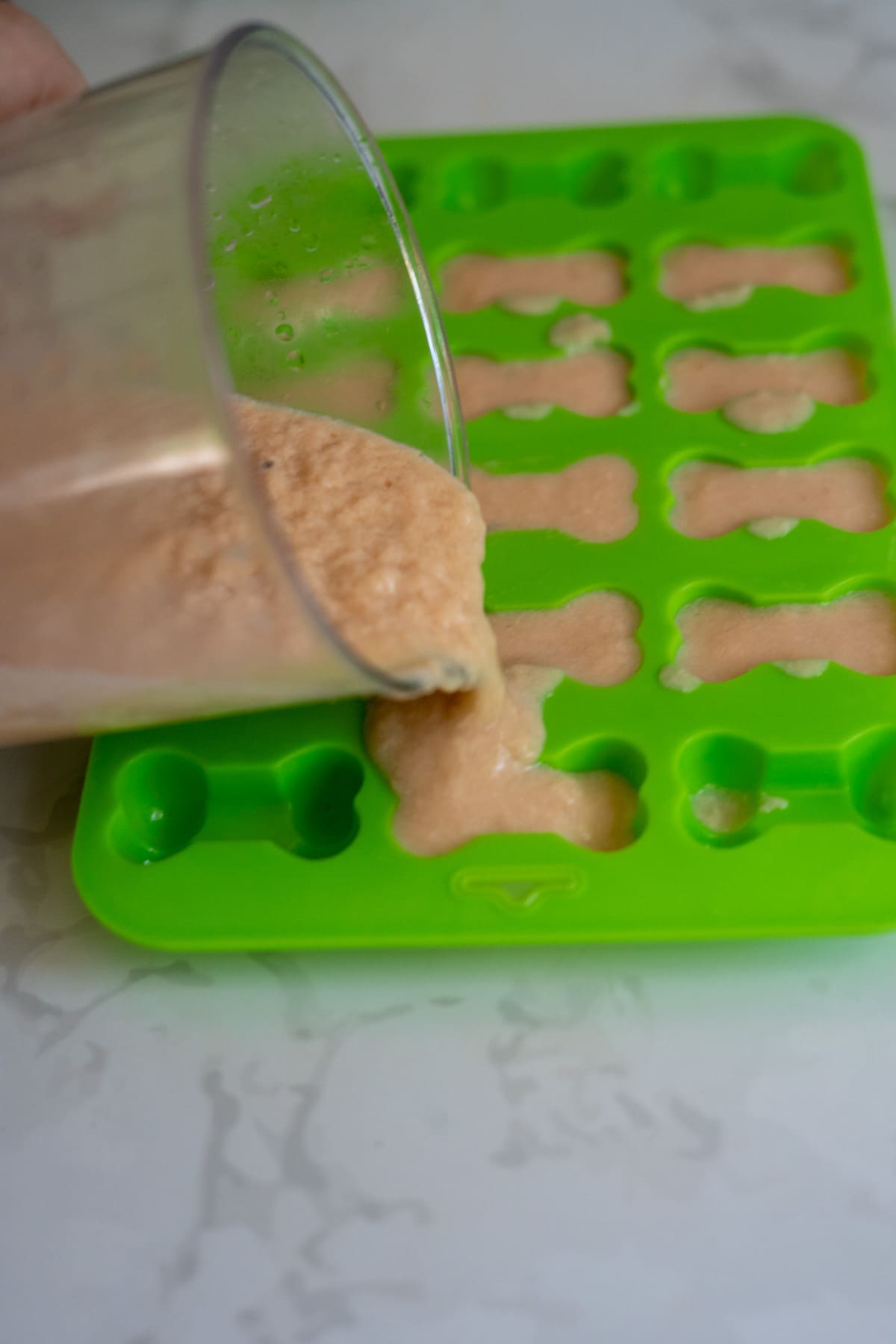 A person pours a beige mixture from a cup into a green silicone mould with bone-shaped cavities on a light-coloured surface, preparing homemade salmon bone broth gummies.