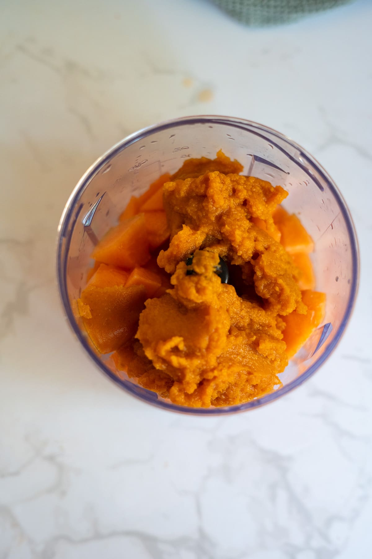 A clear blender jug filled with orange chunks and a thick orange purée, perfect for making Cantaloupe Pumpkin Frozen Dog Treats, sits on a white marble worktop.