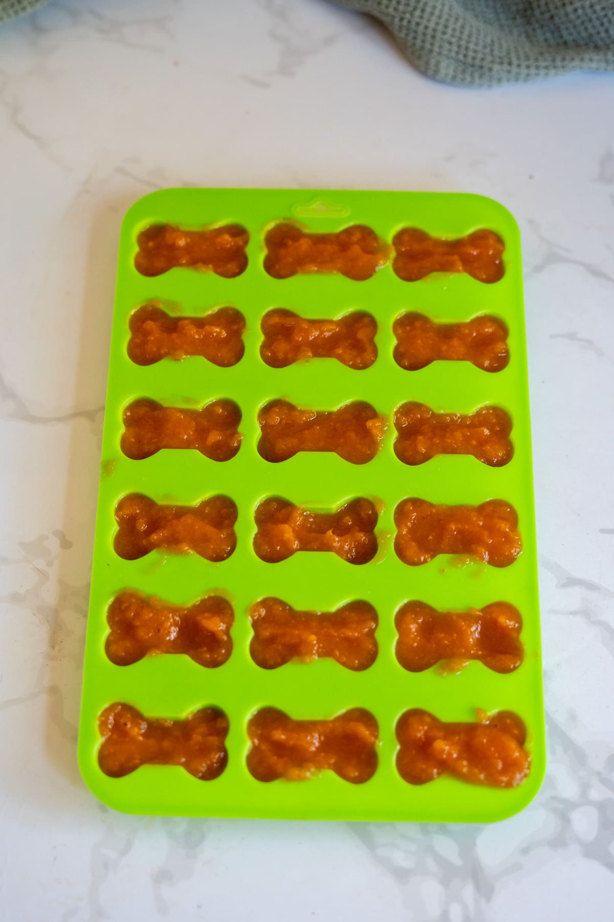 Green silicone mould filled with bone-shaped orange mixture of cantaloupe and pumpkin, creating frozen dog treats, resting on a white marbled surface.