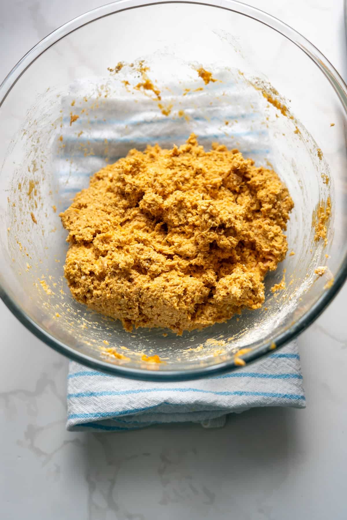 A glass bowl containing a mixture of orange-coloured dough for salmon pumpkin dog biscuits sits on a striped cloth on a white worktop.