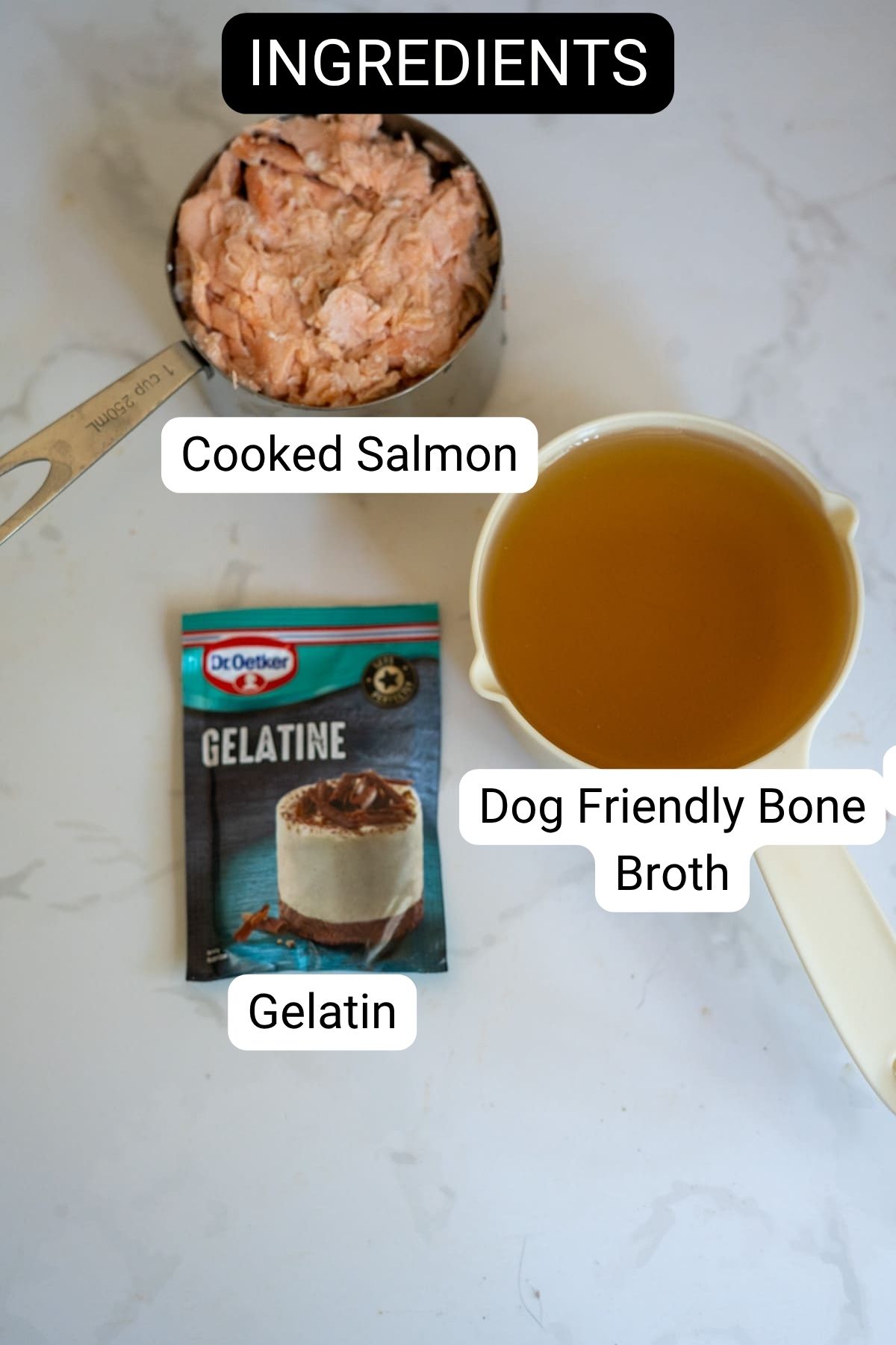 A measuring jug of cooked salmon, a sachet of gelatine, and a jug of dog-friendly bone broth are arranged on a white surface, labelled as ingredients for making salmon bone broth gummies.