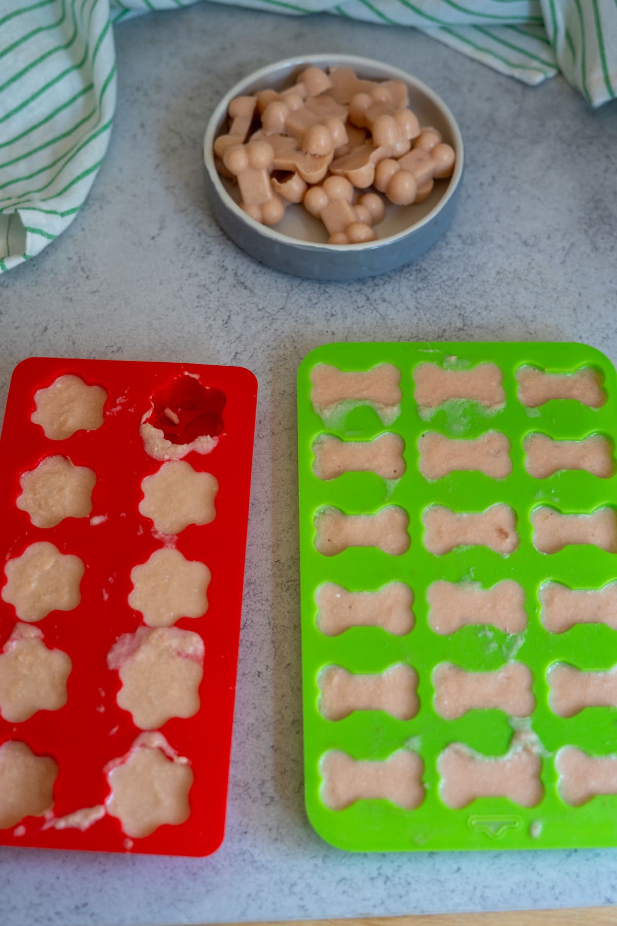 Two silicone moulds filled with light brown batter—one red with paw shapes and one green with bone shapes—are on a counter next to a bowl of finished bone-shaped treats, perfect for creating homemade salmon bone broth gummies.