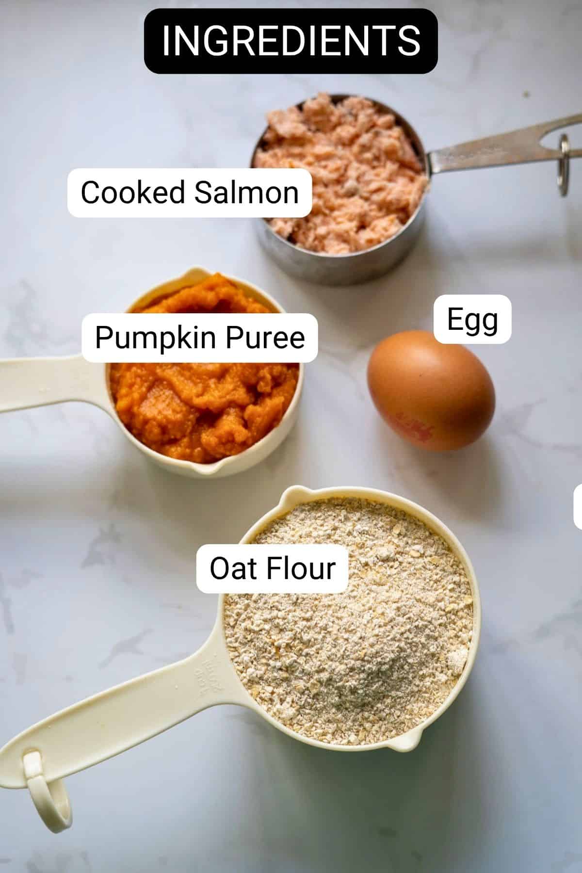 Measuring cups of cooked salmon, pumpkin purée, oat flour, and a whole egg on a marble surface, labelled as ingredients for homemade salmon pumpkin dog biscuits.