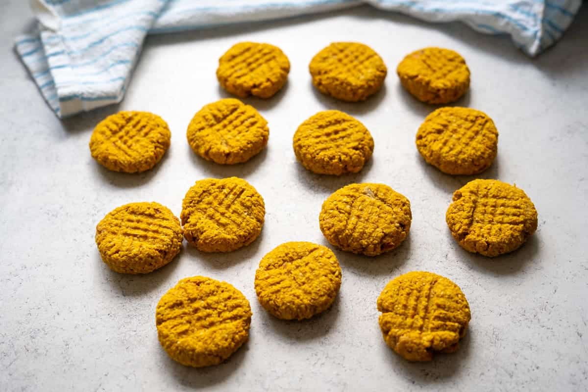 Fourteen round biscuits with a criss-cross pattern on top are arranged on a white surface near a folded striped cloth.