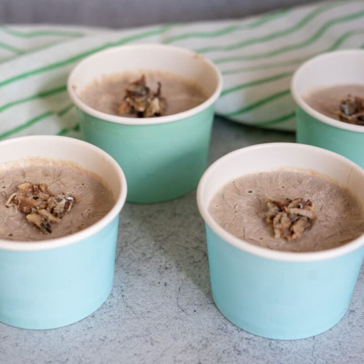 Four light blue paper cups filled with a greyish frozen dessert, each topped with a small cluster of nuts, on a grey surface with a striped cloth in the background.