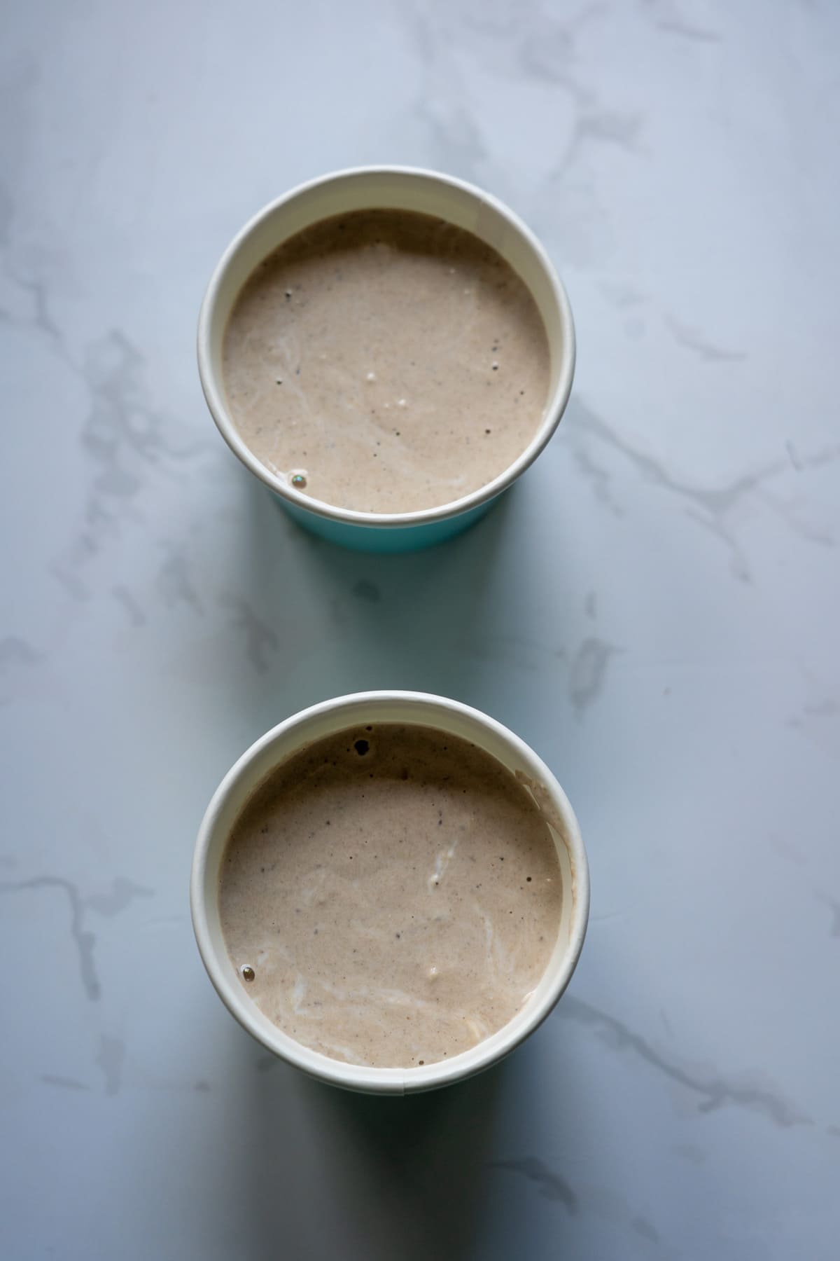 Two paper cups filled with a light brown, creamy sardine ice cream beverage are placed on a white, marbled surface.