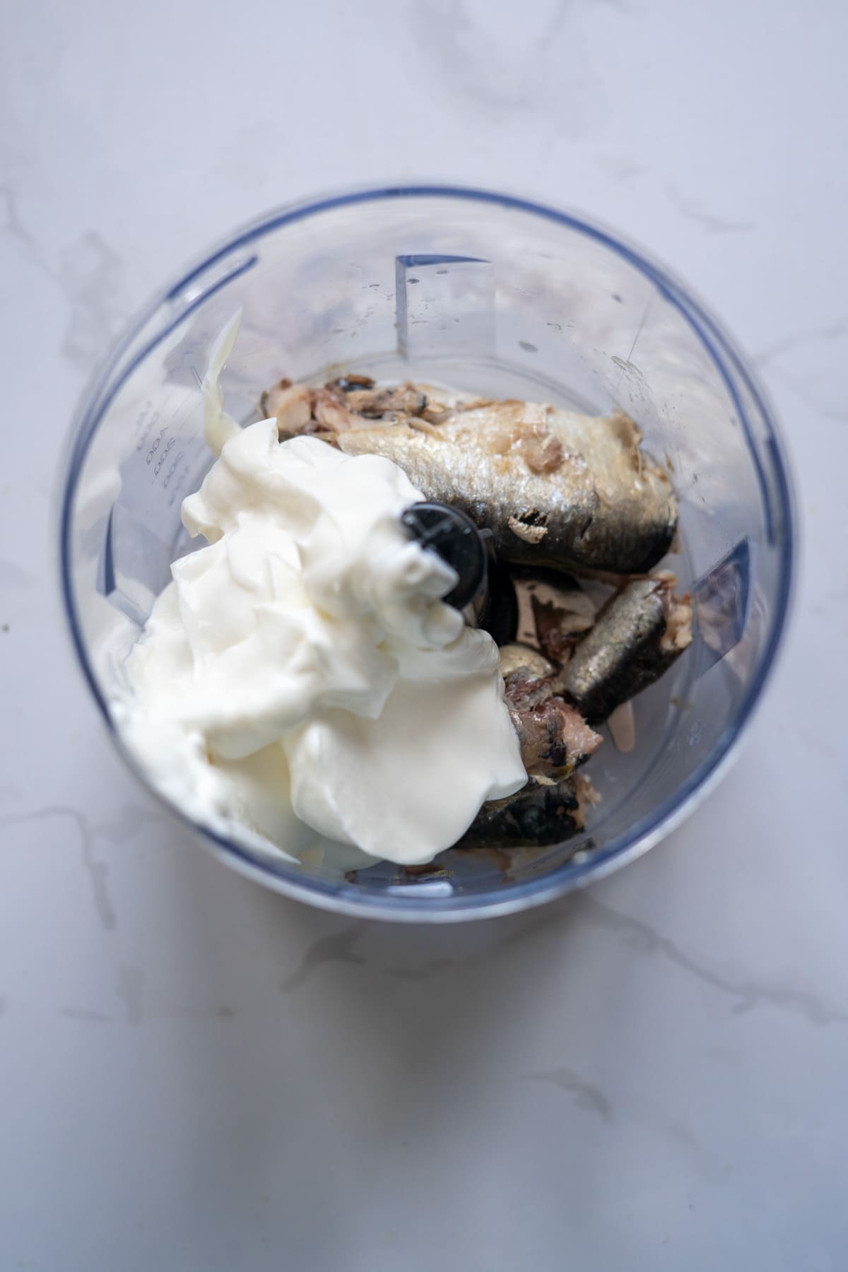 A food processor bowl containing whole sardines and a large dollop of white creamy substance, possibly the base for unique sardine ice cream, rests on a marble worktop.
