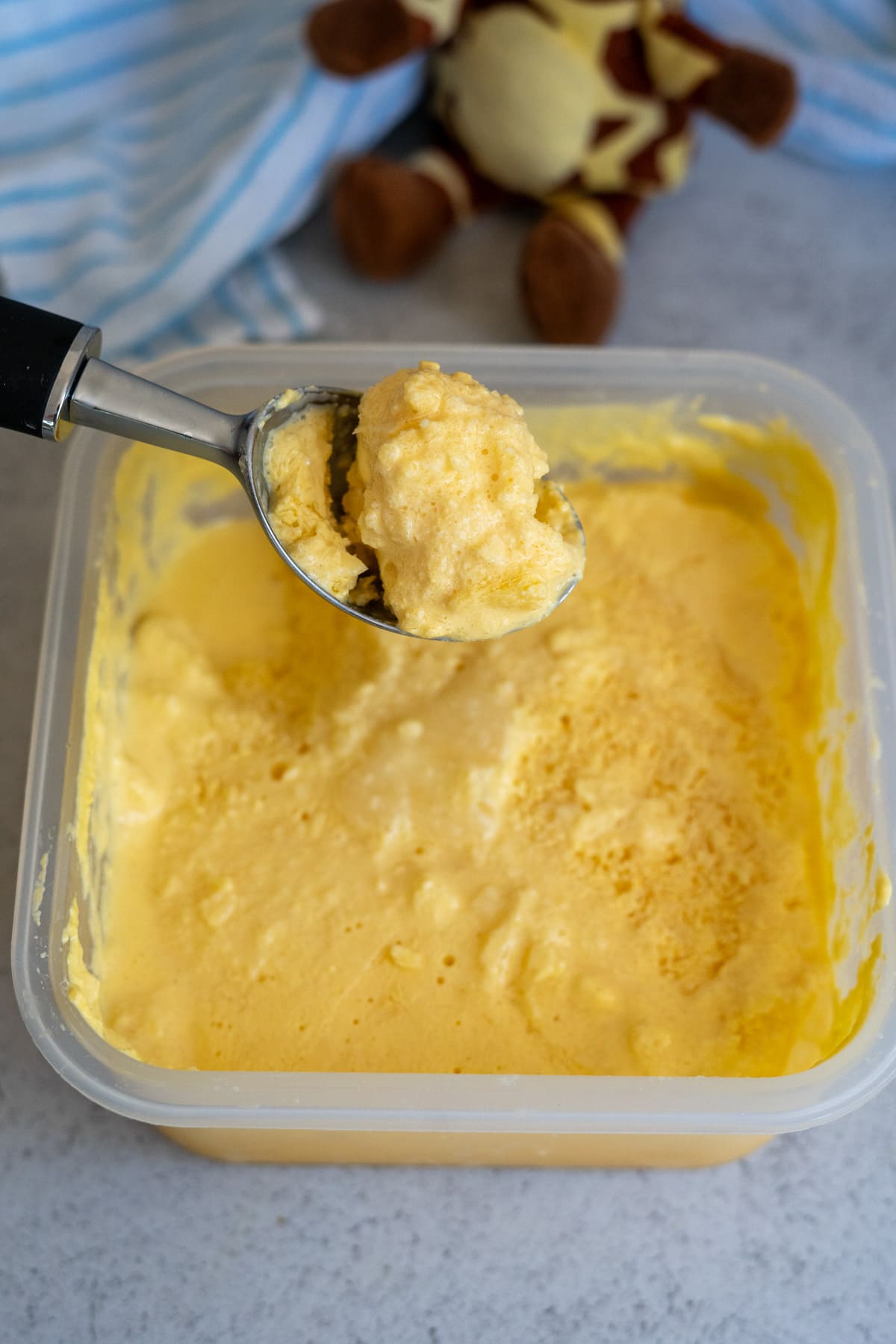 A metal ice cream scoop lifts a serving of yellow ice cream from a plastic tub, with a soft toy and striped cloth in the background.