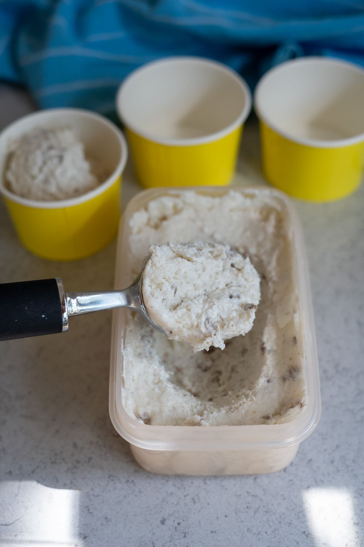 A metal ice cream scoop holds a portion of beef dog ice cream above a rectangular container, with three yellow paper cups—one partially filled—nearby on a worktop.