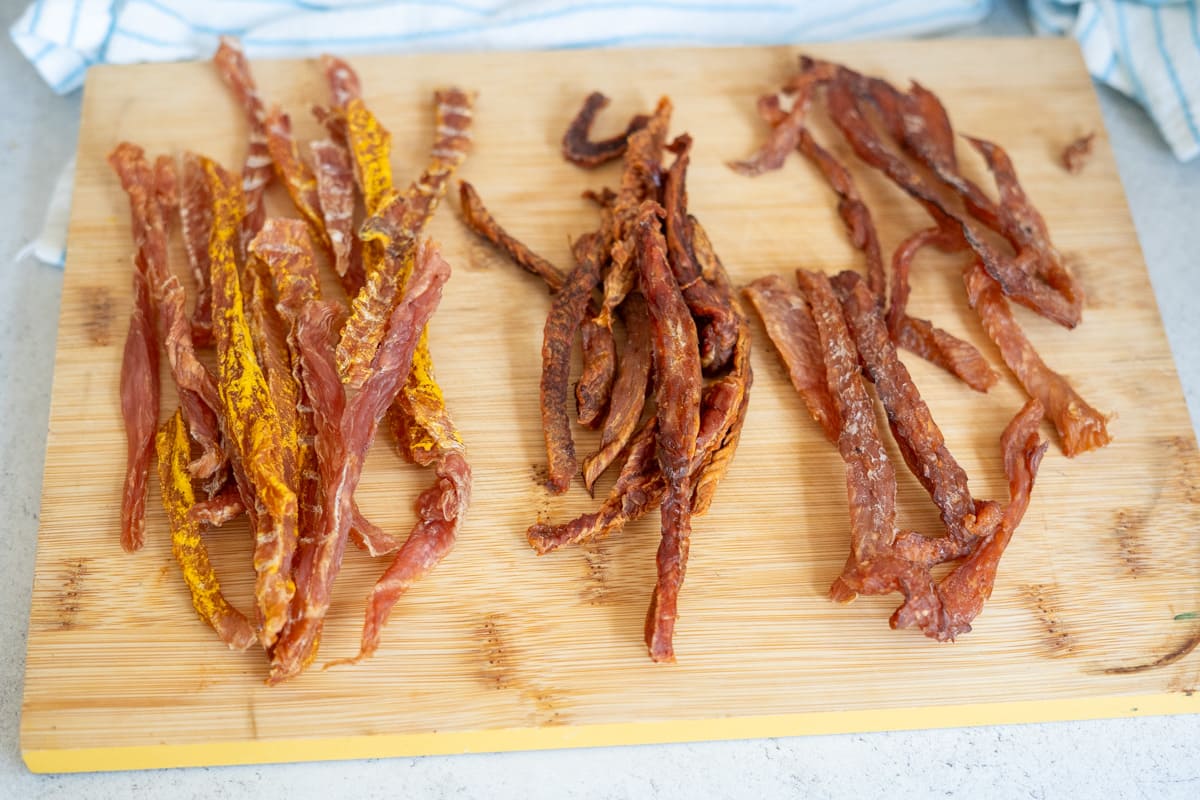 Three types of homemade jerky strips, including turkey jerky for dogs, are arranged on a wooden chopping board, with a striped cloth partially visible in the background.